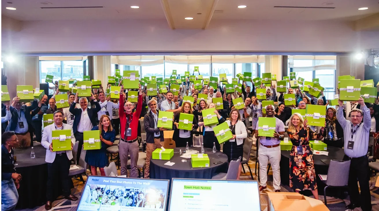 Group of Rocket Software employees standing in a conference room holding green signs, smiling and facing the camera, with screens and desks visible in the foreground.